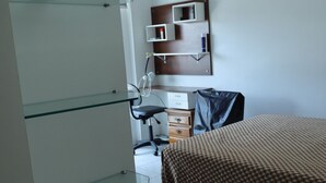 Hotellihuone, jossa on sänky, työpöytä, tuoli, riippuvalaisin ja lasihyllyillä varustettu vaatekaappi.