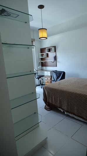 A hotel room with a bed, a desk, a chair, a hanging lamp, and a wardrobe with glass shelves. - Ótimo Apto à 650m da praia do Forte em Cabo Frio (Cabo Frio)
