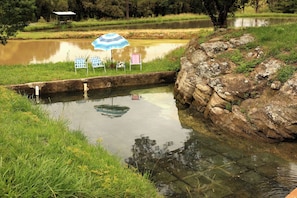 A tranquil outdoor setting with a small pond, a stone embankment, and three chairs under a striped umbrella.