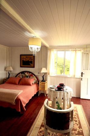 A bedroom with a bed, two bedside lamps, a chandelier, a window with curtains, a small table with a lace tablecloth, and a framed picture on the wall.