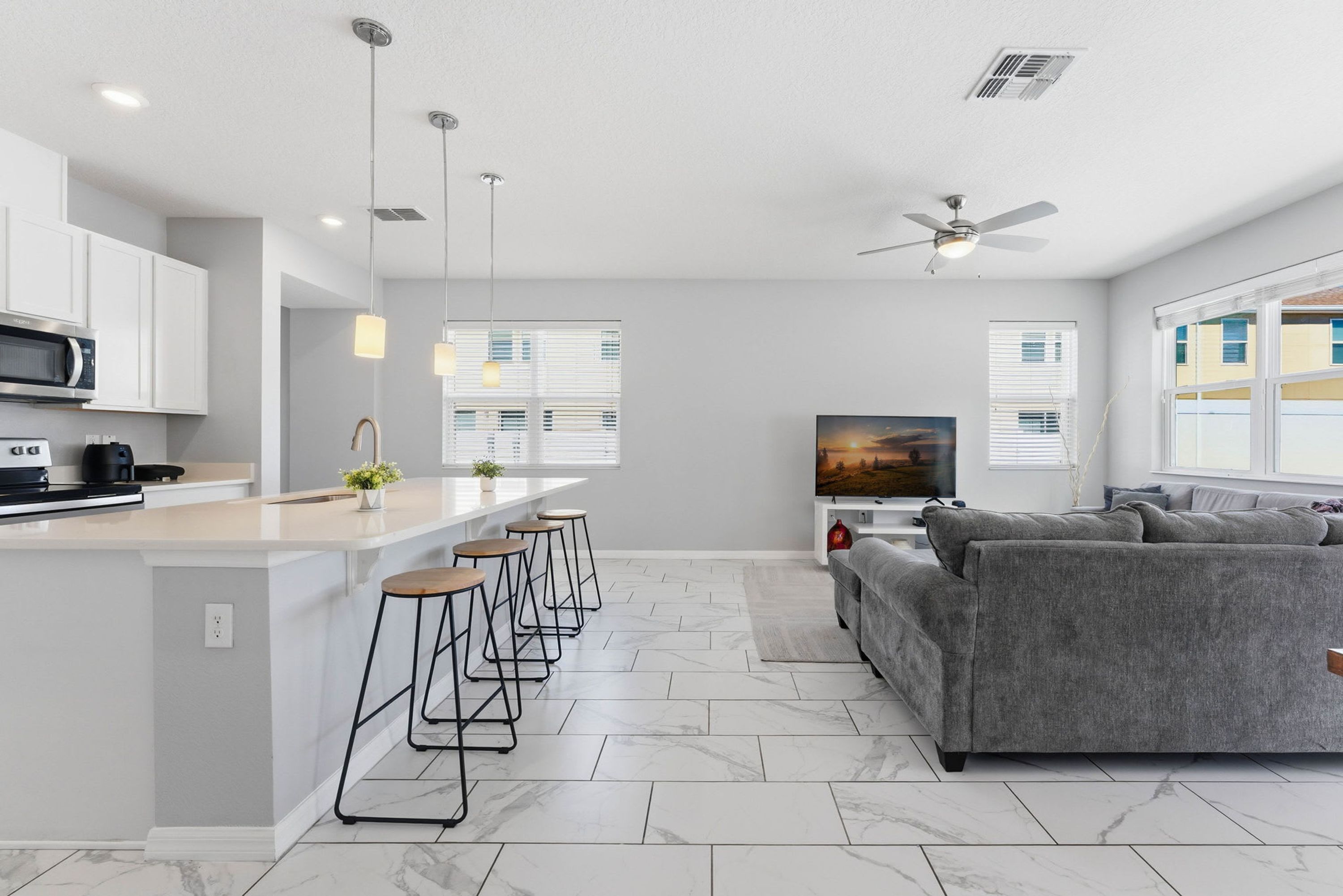 A modern kitchen with a central island, bar stools, a grey sofa, and a TV in a living area.