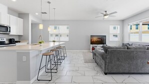 Una cucina moderna con isola centrale, sgabelli da bar, un divano grigio e una TV nella zona living.