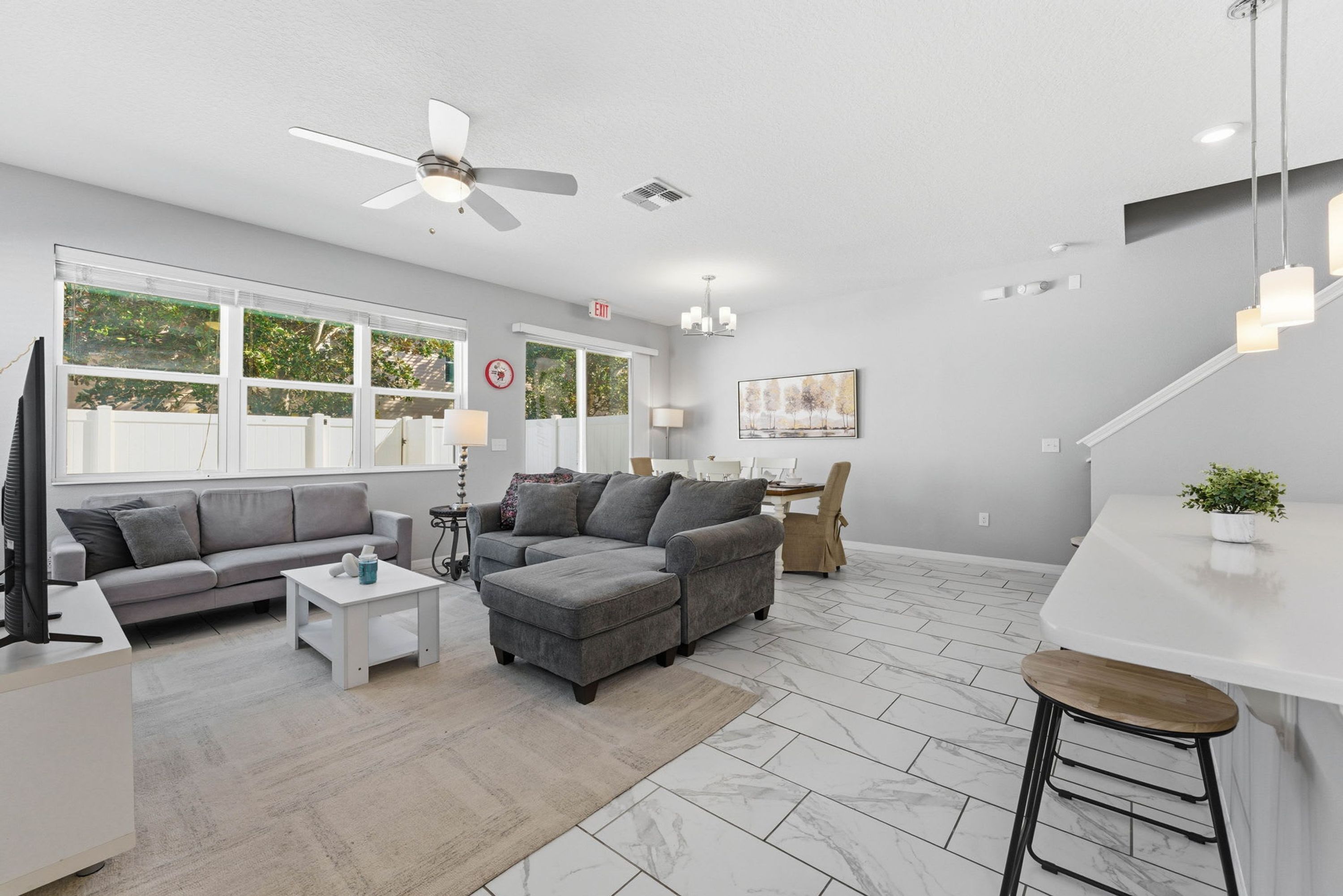 A modern living room with a grey sofa, a white coffee table, and a small rug. There is a ceiling fan, a painting on the wall, and a bar area with stools.