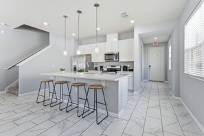 Een moderne keuken met witte kasten, een centraal kookeiland met krukken, roestvrijstalen apparatuur en hanglampen.