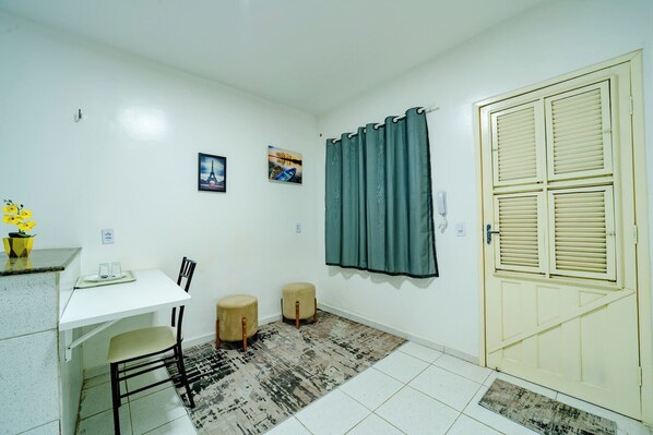 Una habitación pequeña y limpia con un escritorio blanco, dos sillas, un tapete pequeño, una cortina verde y una puerta con contraventanas.