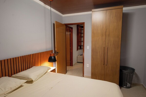 A bedroom with a bed, a wooden wardrobe, a hanging lamp, and a trash bin.