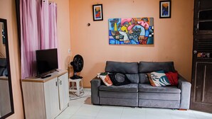 A living room with a grey sofa, a flat-screen TV, a fan, and two paintings on the wall. - Casa bem equipada com WiFi e ótima localização (Belém)