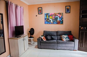 Ein Wohnzimmer mit grauem Sofa, einem Flachbildfernseher, einem Ventilator und zwei Bildern an der Wand.
