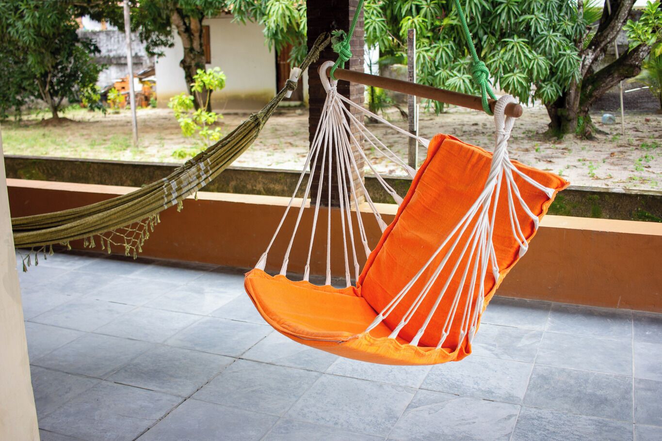 Una silla hamaca colgada con cuerdas, unida a una viga de madera, en un espacio exterior con suelo de baldosas y follaje al fondo.