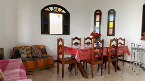Zona pranzo con un tavolo apparecchiato per quattro, circondato da sedie in legno. Sopra il tavolo si vedono due vetrate istoriate, e a sinistra una poltrona con una coperta colorata.