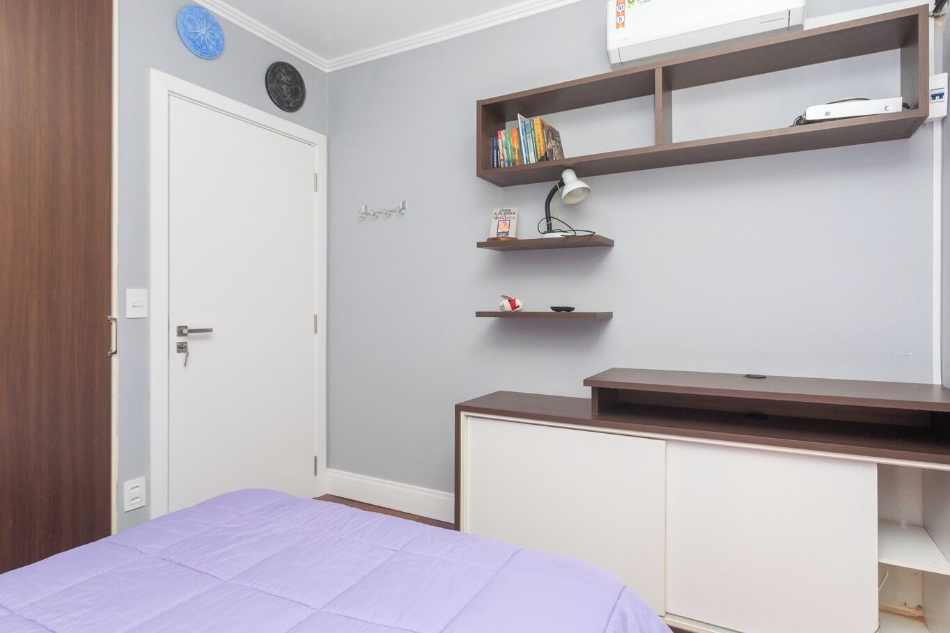 A bedroom with a bed, a desk, shelves with books, a lamp, and a wardrobe.