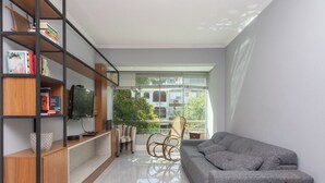 Una sala de estar moderna con un sofá gris, una estantería de madera, un televisor y vistas a zonas verdes.