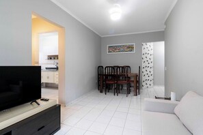 A modern living room with a flat-screen TV, a dining area with a table and chairs, and a white sofa. - Apto com excelente localização a 50m da praia (Praia Grande)