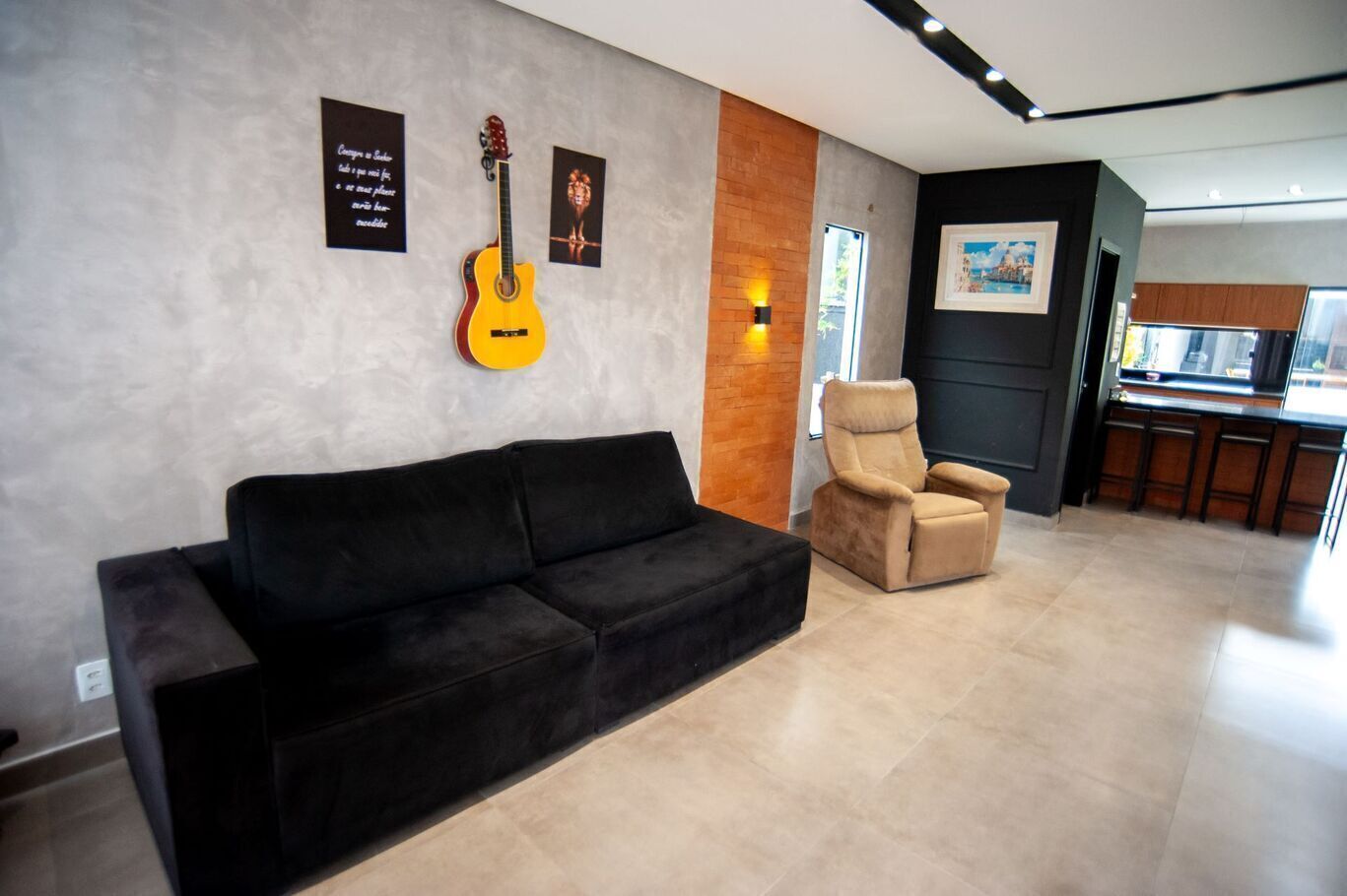 A modern living room with a black sofa, a beige armchair, a guitar, and a wall-mounted light fixture.