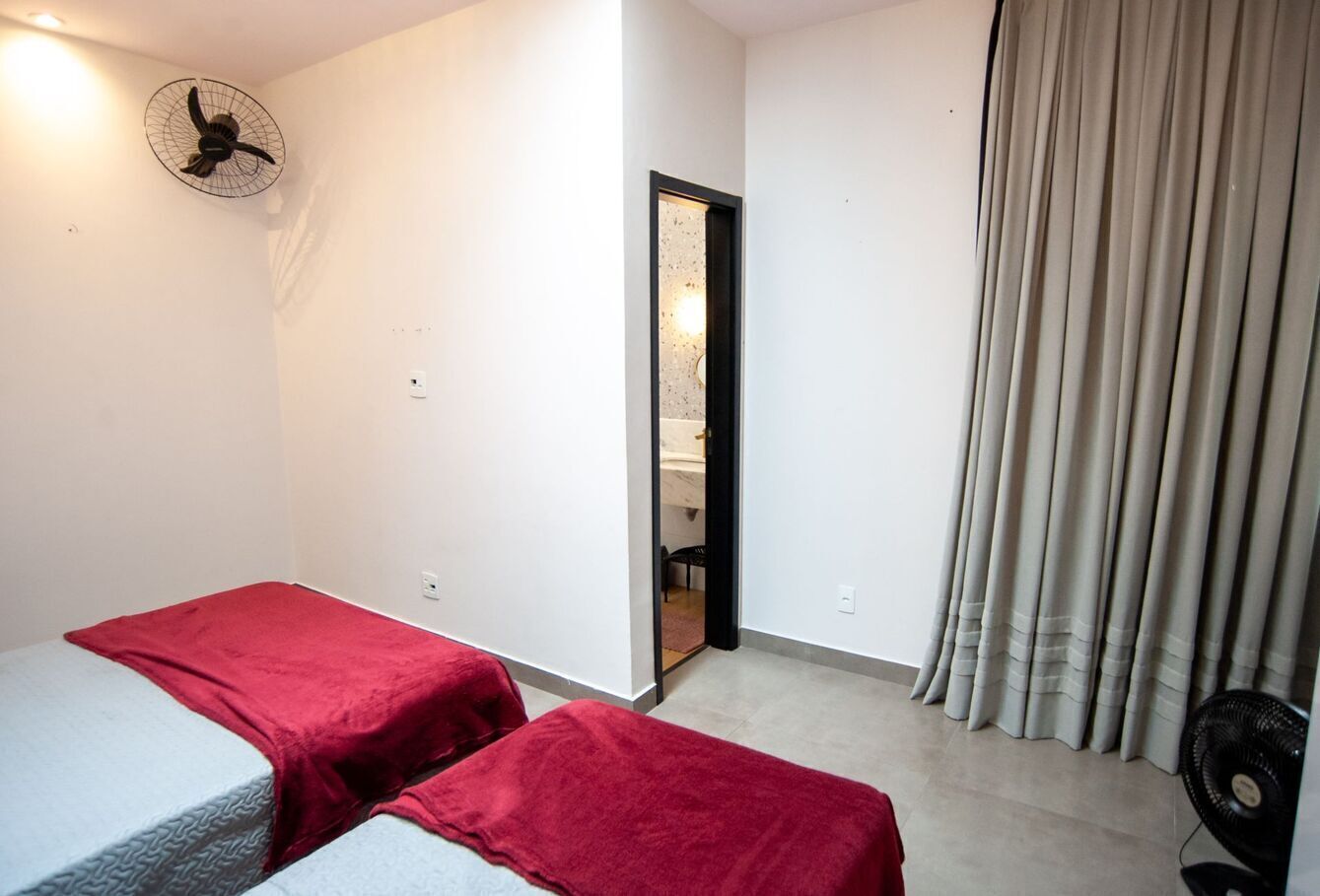 Una habitación con dos camas, ventilador de techo, puerta que da a un baño y ventana con cortinas.