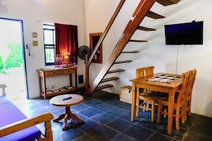 Ein gemütliches Zimmer mit einer Holztreppe, einem Esstisch mit Stühlen, einem kleinen runden Tisch mit einem Sessel, einem an der Wand befestigten Fernseher und einem Fenster mit Vorhängen.