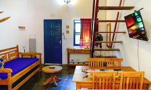 Ein gemütliches Wohnzimmer mit blauer Tür, Holzmöbeln, einem kleinen Tisch und einem an der Wand montierten Fernseher.