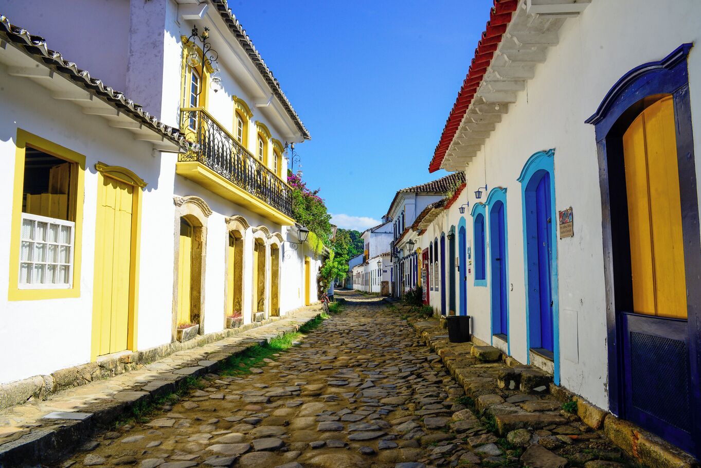 A cobblestone street flanked by buildings with yellow and blue doors, under a clear blue sky.