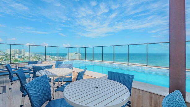Ein Poolbereich auf dem Dach mit blauen Korbstühlen und Tischen mit Blick auf eine Stadtlandschaft und das Meer.