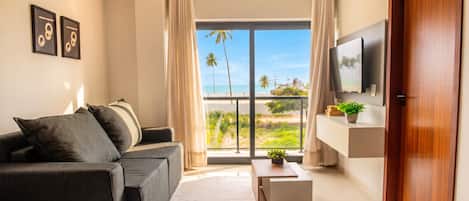 A modern living room with a sofa, a coffee table, a flat-screen TV, and a view of the beach.