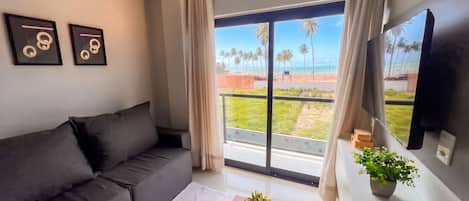 A modern living room with a black sofa, a small table with a plant, and a balcony with a view of palm trees and the beach.
