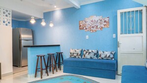Una sala de estar con un sofá azul, tres taburetes de madera, un refrigerador y una puerta con barandales decorativos.