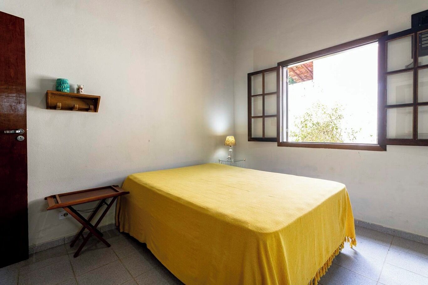 Une pièce avec un lit recouvert d’un tissu jaune, un banc en bois, une petite étagère avec quelques objets, et une fenêtre donnant sur un paysage.