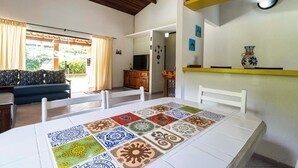 Un coin repas avec une table recouverte de nappes à motifs, un canapé avec des coussins, un téléviseur et un espace cuisine avec une étagère jaune.