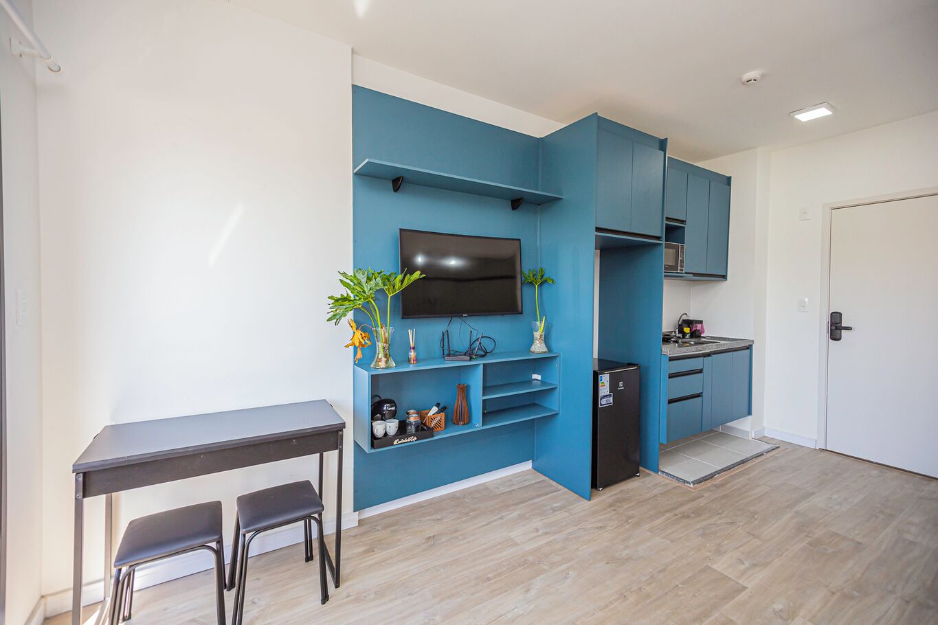 Um espaço compacto com cozinha compacta, área de refeições com mesa e cadeiras, e televisão fixada na parede azul.