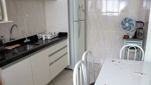 Una cocina con armarios blancos, encimera negra, lavabo, refrigerador, ventilador y mesa de comedor con sillas.