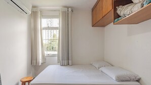 Une petite pièce propre, avec un lit, une fenêtre avec des rideaux, une étagère en bois où sont pliés des vêtements et un petit tabouret.