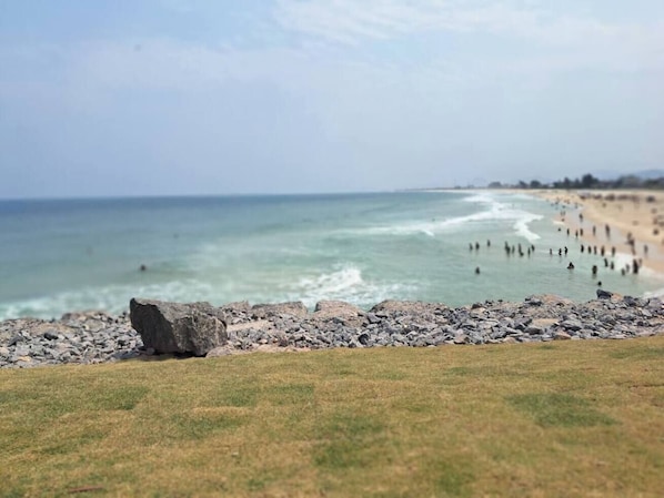 Une plage bordée d’une côte rocheuse, avec un tapis d’herbe au premier plan et une vue lointaine sur l’océan.