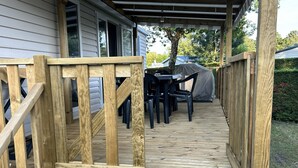 Eine Holzterrasse mit überdachtem Tisch und Stühlen, einem überdachten Grill und Blick auf ein Haus im Hintergrund.