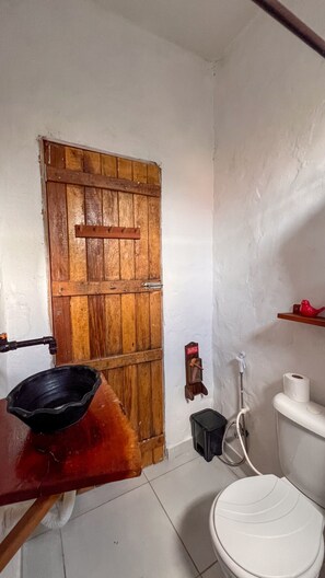 A small, compact bathroom with a wooden door, a toilet, a small shelf with a red object, and a black basin on a wooden table. - Casa Brisa das Águas: Pool and BBQ (Tatuamunha)