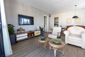 A modern living room with a sofa, chairs, a coffee table, a TV, and a staircase leading to another room. - Cobertura Duplex com piscina a 200m da Praia (Guarujá)