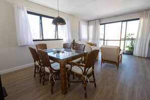 A spacious living room with a glass-top dining table surrounded by wicker chairs, a sofa, and a large window with curtains. - Cobertura Duplex com piscina a 200m da Praia (Guarujá)