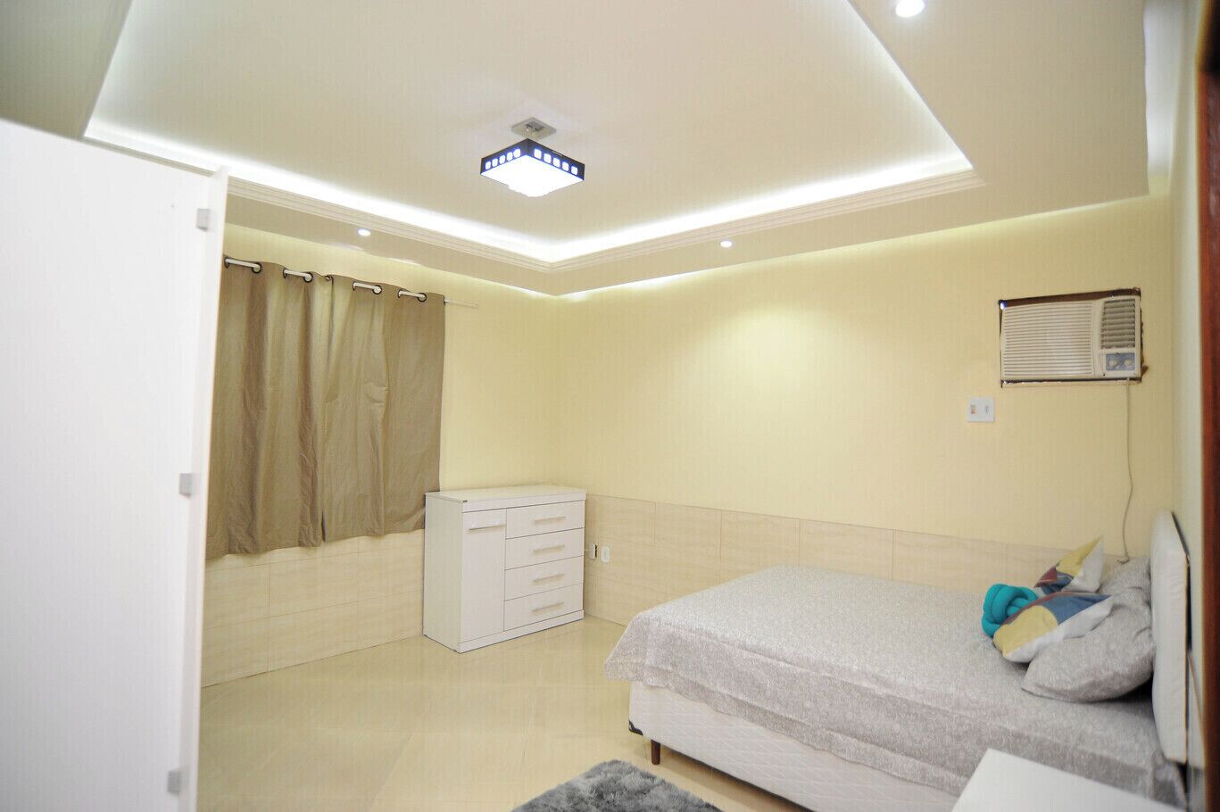A bedroom with a bed, a dresser, a window with curtains, and a ceiling light.