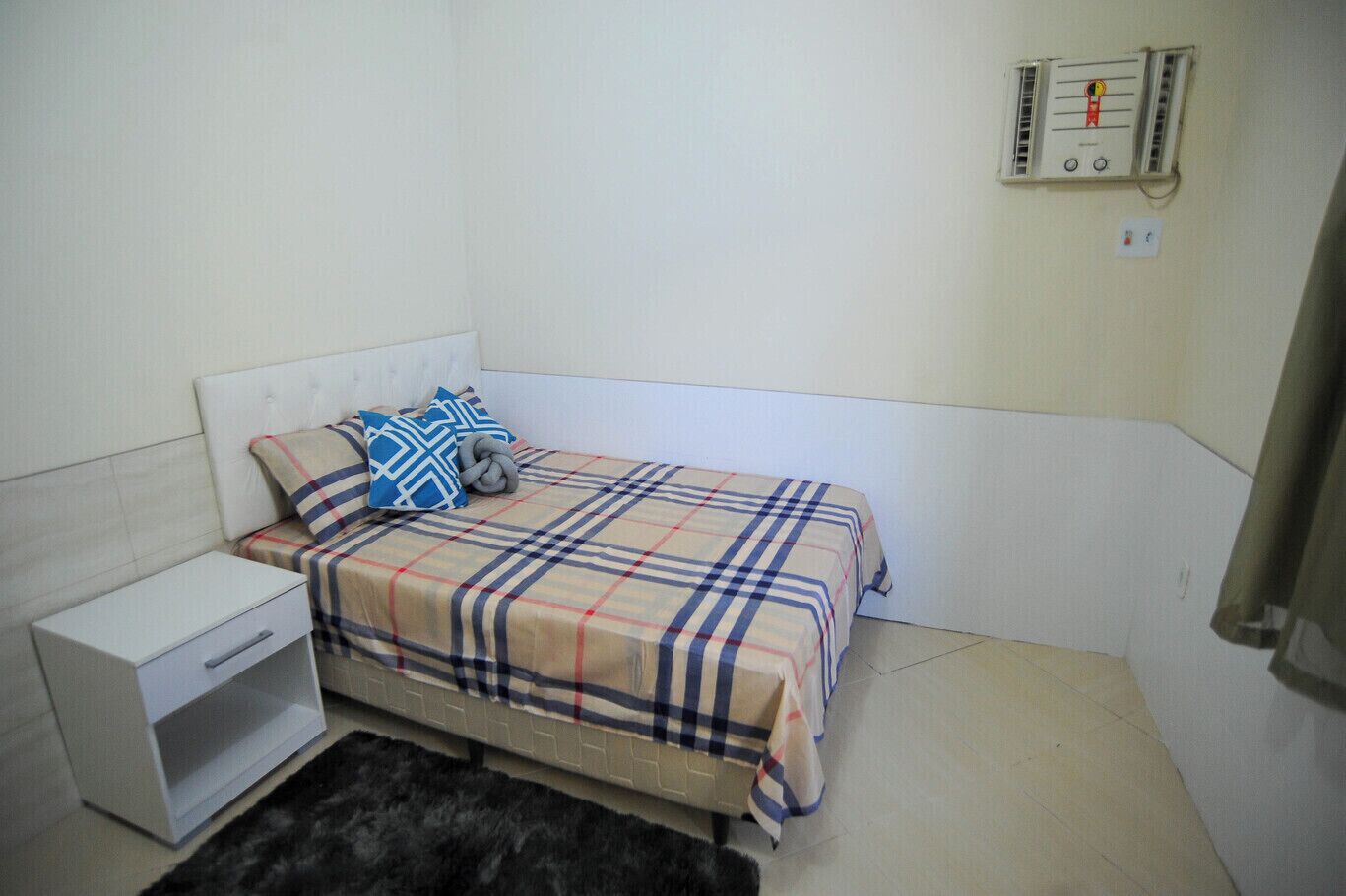 Een kleine kamer met een eenpersoonsbed, een wit hoofdeinde, een nachtkastje en een stopcontact aan de muur.