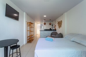 Ein Schlafzimmer mit Bett, einem kleinen Tisch und einem Sessel. Im Hintergrund befindet sich eine Küchenzeile mit einer Mikrowelle, einem Spülbecken und einem Herd.