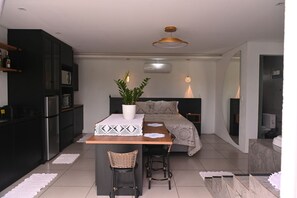 Eine moderne Küche mit dunklen Schränken, einem Kühlschrank und einer Mikrowelle. Es gibt einen Essbereich mit einem Tisch, Stühlen und einem Blumentopf. Der Raum verfügt über ein Bett mit Kopfteil und einen Nachttisch.