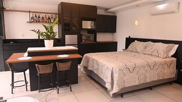 Quarto compacto com cama, pequena cozinha e área de refeições para dois.