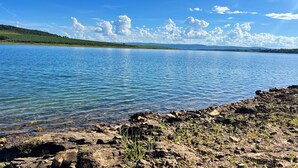 A lake with clear water, a rocky shoreline, and a distant green hill under a blue sky with clouds.