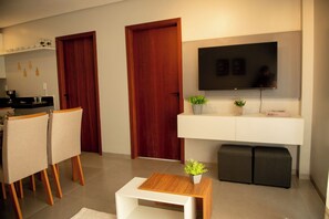 Un salon moderne avec un téléviseur, un coin repas avec des chaises, une petite table avec une plante, et un coin cuisine avec des placards et un plan de travail.