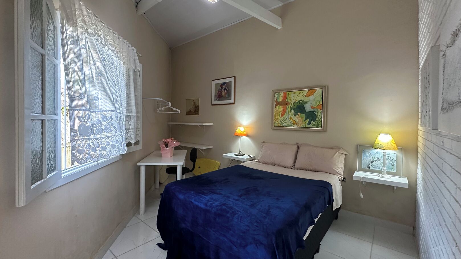 Pequeña habitación con cama, escritorio, silla, lámpara colgante y estante con lámpara.