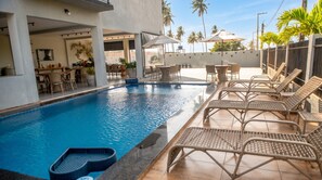 An indoor swimming pool with lounge chairs and tables, surrounded by a covered patio area with umbrellas. - Beachfront apt: balcony, pool, pet friendly (Japaratinga)