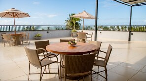 A round wooden table with wicker chairs and two umbrellas on a balcony with a view of the ocean. - Beachfront apt: balcony, pool, pet friendly (Japaratinga)