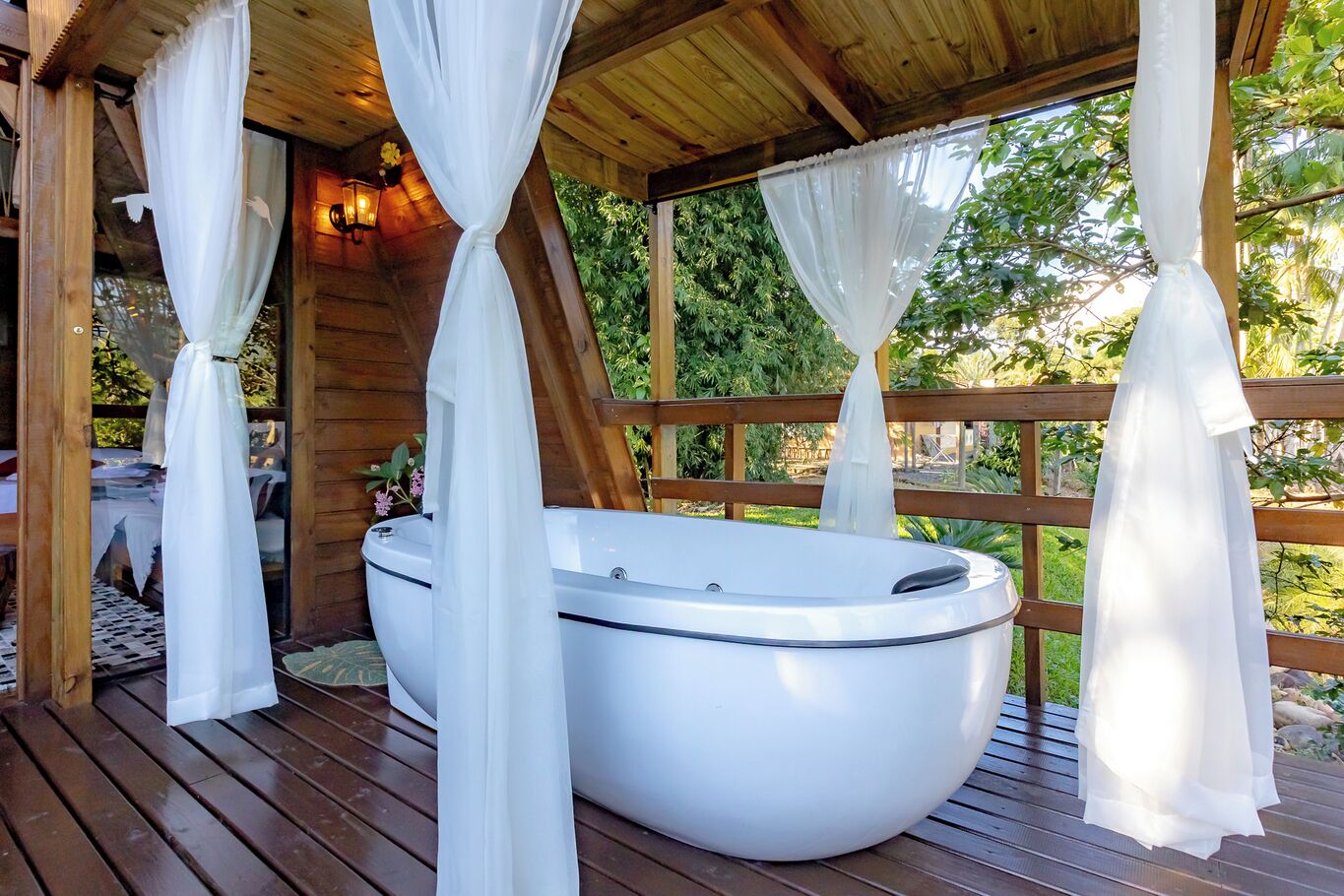 Un bain à remous installé dans un gazebo en bois, agrémenté de rideaux blancs et entouré de verdure.