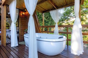 A hot tub in a wooden gazebo with white curtains, surrounded by greenery.