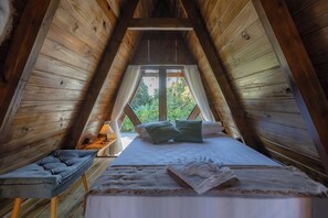 A cozy attic bedroom with a wooden sloped ceiling, a bed with pillows, a bedside table with a lamp, and a view of greenery outside.