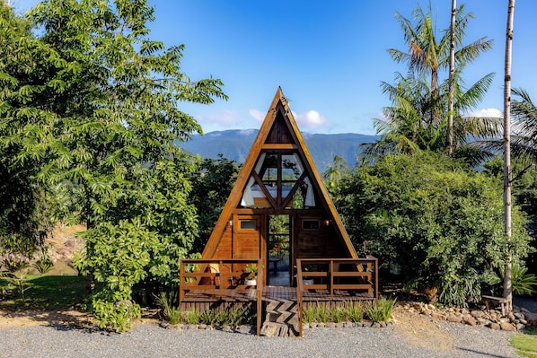 A wooden triangular cabin with a balcony, surrounded by lush greenery and mountains in the background.